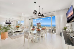 A modern living room with a glass dining table, white sofas, and a large TV screen.