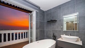 A modern bathroom with a large bathtub, a vanity with a mirror, and a view of a sunset through an open door. - Merci Sunset Town Phu Quoc (Phu Quoc Island)