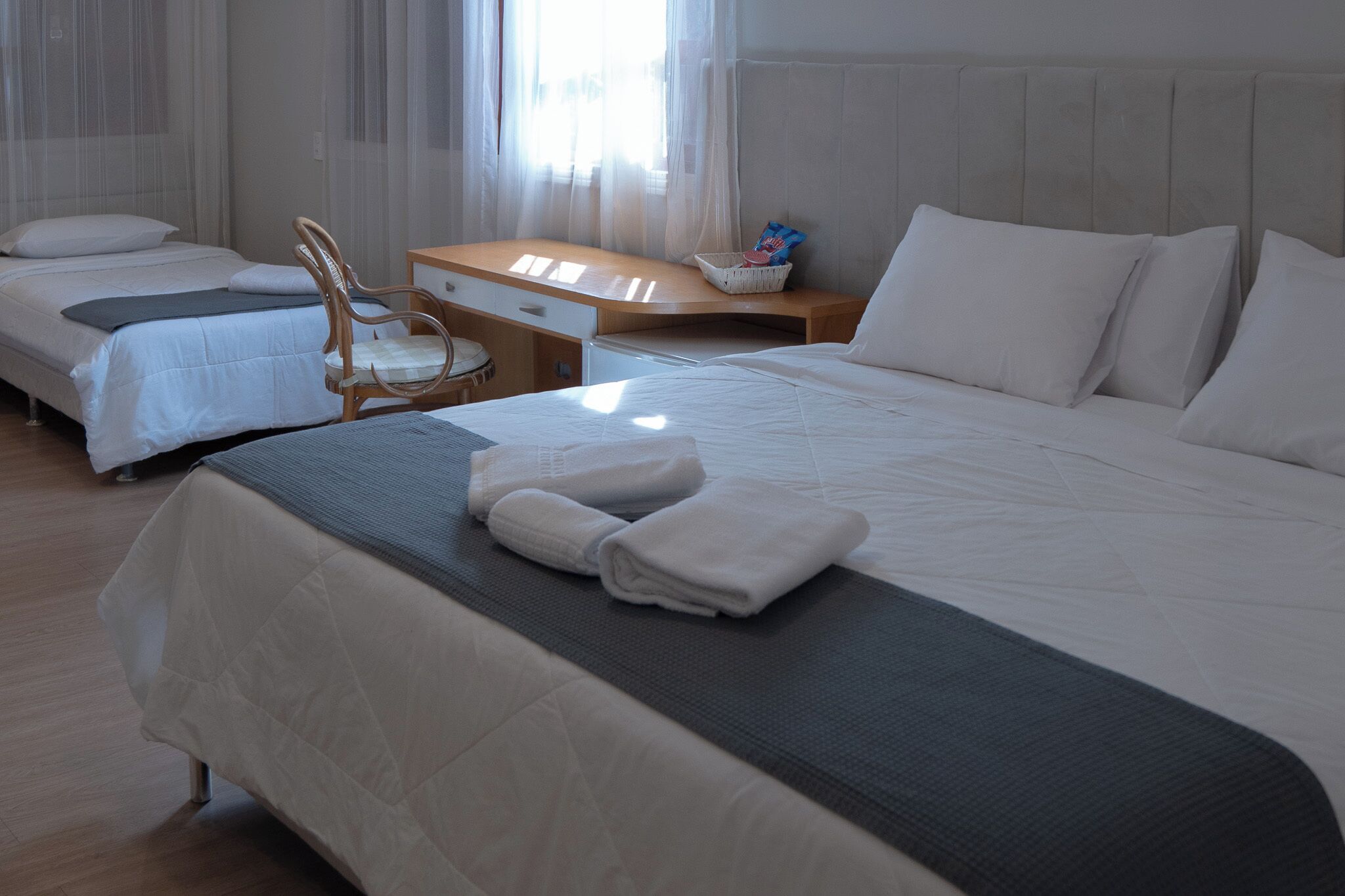 A hotel room with two beds, a desk, a chair, and a window with sheer curtains.