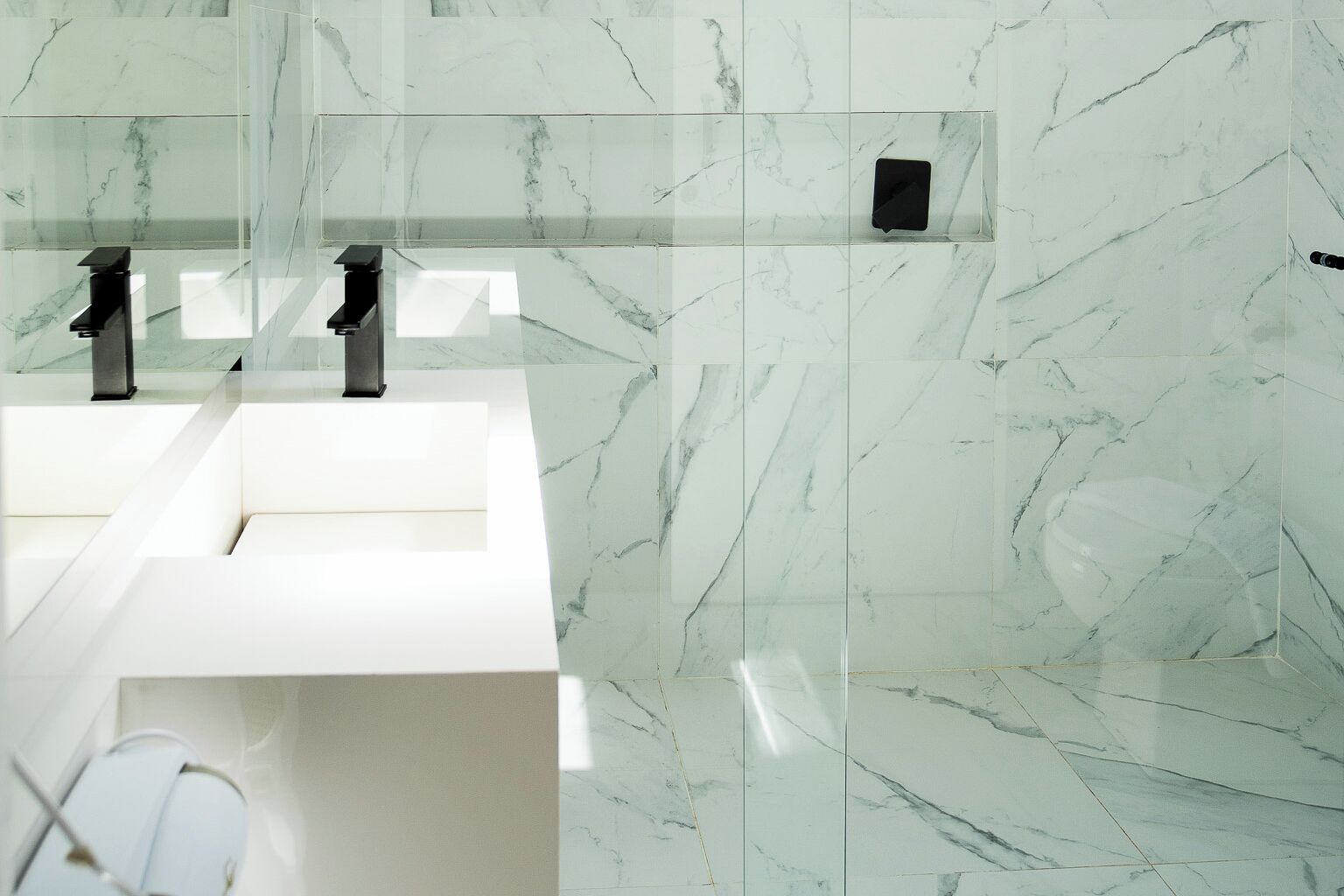 A modern bathroom with a white marble sink, two faucets, and a glass partition.