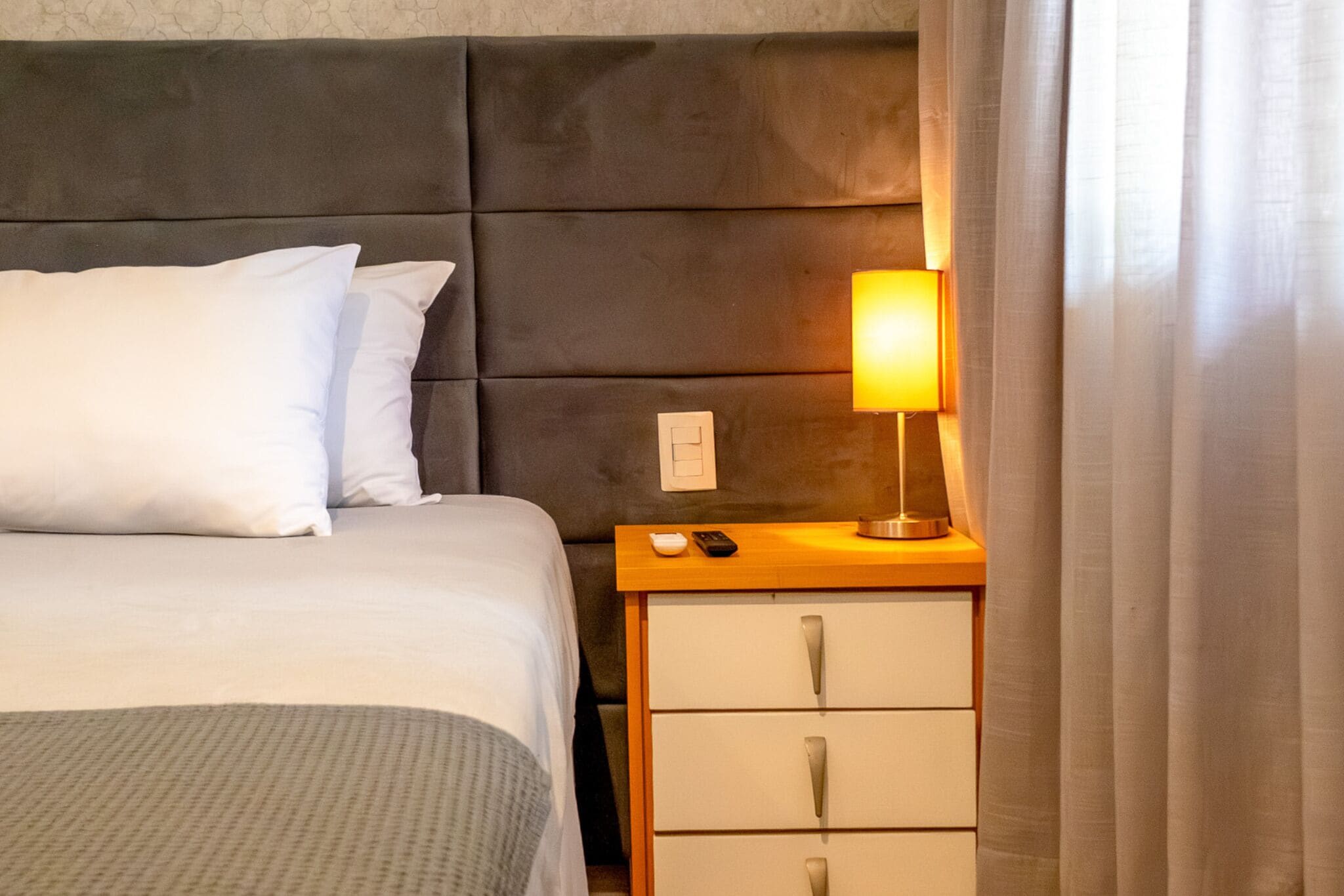 Una cama bien tendida con ropa blanca, una mesita de noche con una lámpara y un interruptor de luz en la pared.