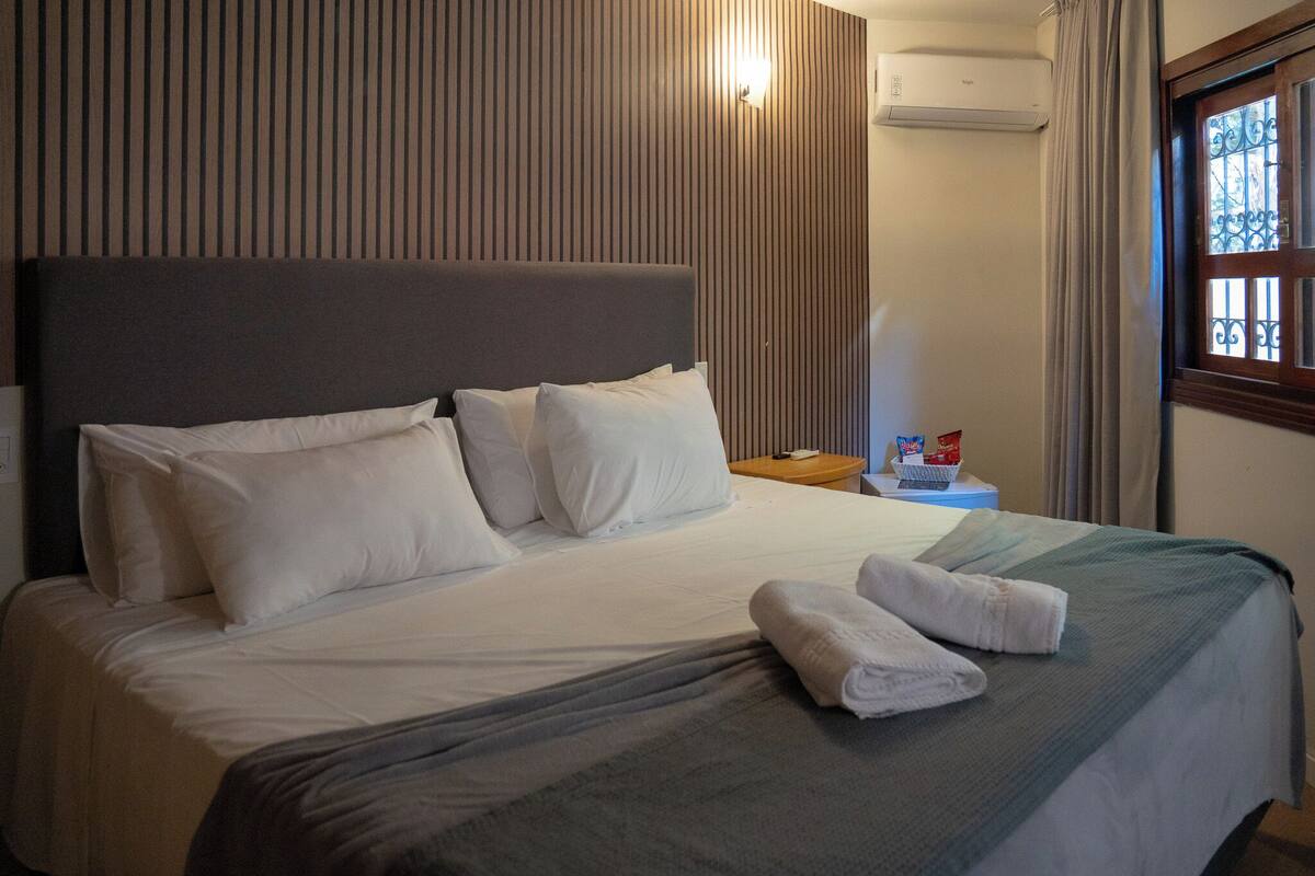Une chambre d’hôtel avec un lit, des oreillers, une table de chevet, un panier et une fenêtre avec des rideaux.