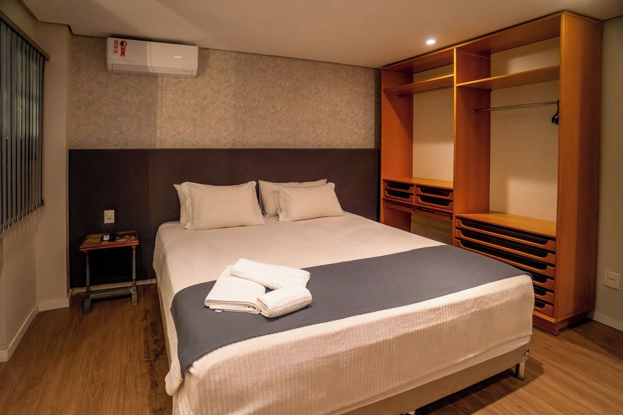 A neatly made bed with pillows, a bedside table, an air conditioner, and a wooden wardrobe.