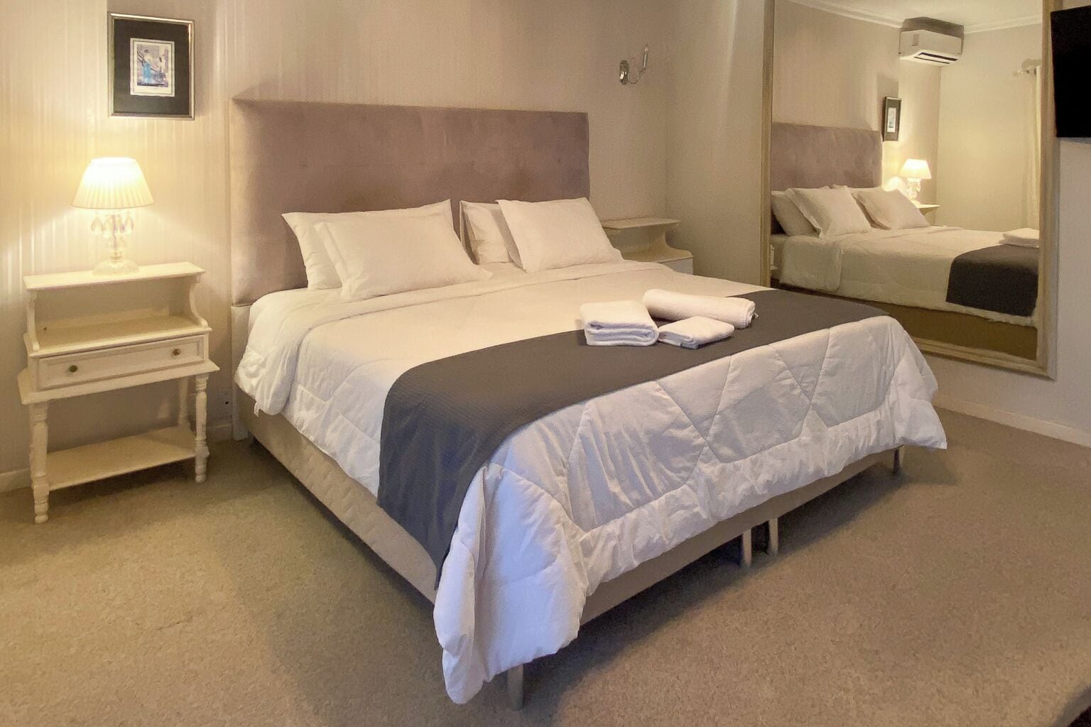 A neatly made bed with a headboard, a bedside table with a lamp, and a mirror on the wall.