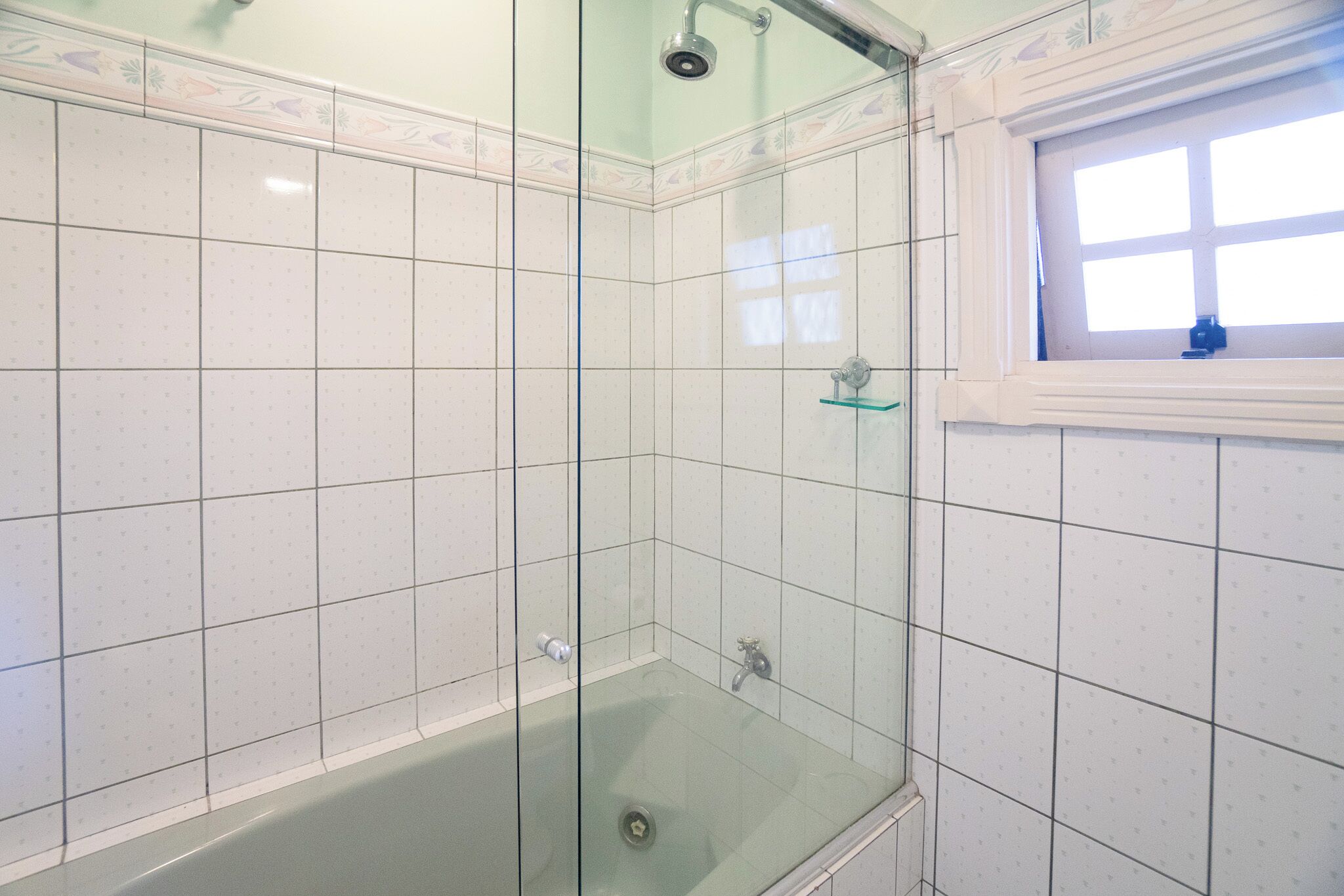 A glass-enclosed shower with a window, white tiled walls, and a bathtub.