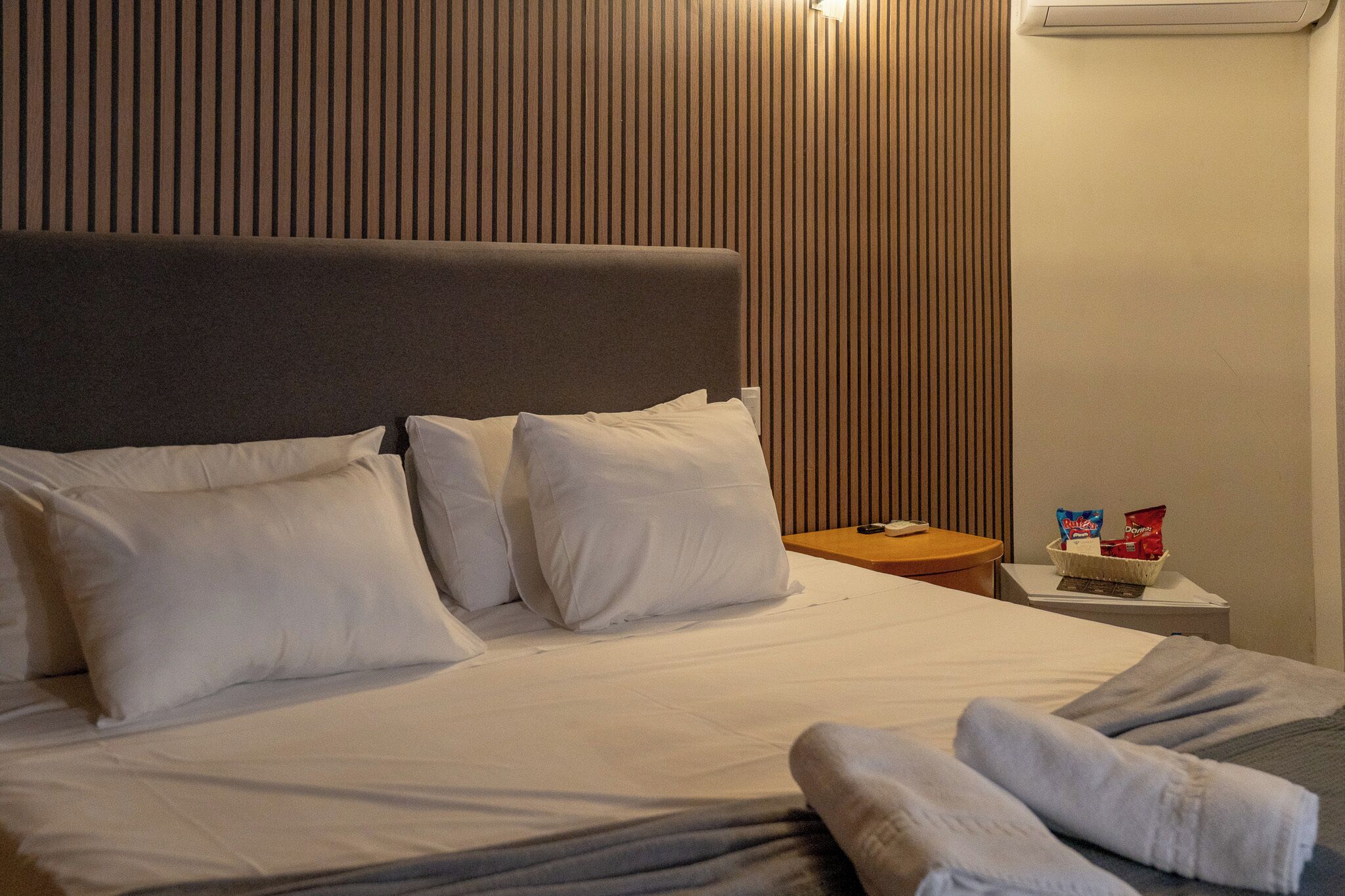 Una cama bien hecha con sábanas blancas, un cabecero gris y una mesita de noche con una cesta que contiene aperitivos.