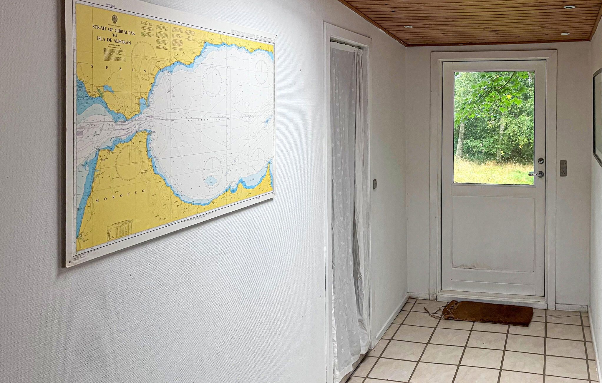 Un couloir avec une carte accrochée au mur, une porte blanche et une fenêtre donnant sur un paysage verdoyant.