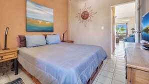 A bedroom with a bed, nightstand, and a painting of a sunset. - Viva Condominiums Viva 28 (San José del Cabo)