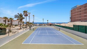 A tennis court with a net, surrounded by palm trees and a building with multiple balconies. - Viva Condominiums Viva 28 (San José del Cabo)