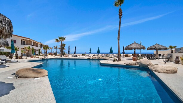 A clear blue pool with lounge chairs and umbrellas, surrounded by palm trees and a building with a view of the ocean. - Viva Condominiums Viva 28 (San José del Cabo)