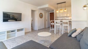 A modern living room with a grey sofa, a TV mounted on the wall, and a small white table. There is a kitchen area with white cabinets and a dining table with chairs. - Cozy apartment in Fuengirola (Fuengirola)