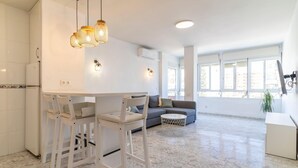 A modern kitchen with white cabinetry, a dining area with white chairs, a grey sofa, and a large window providing natural light. - Cozy apartment in Fuengirola (Fuengirola)