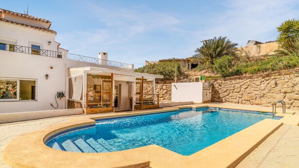 Une piscine à bord incurvé, un patio couvert et un mur de pierre en arrière-plan.