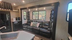 A compact living space with a kitchenette, a leather sofa, and a window providing a view of greenery. - 42- foot long Comfortable Camper near Marble Falls! (Marble Falls)