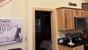Avoin, loft-tyylinen huone, jossa on puinen portaikko, keittiöalue puisilla kaapeilla ja mikroaaltouuni sekä taulu seinällä.