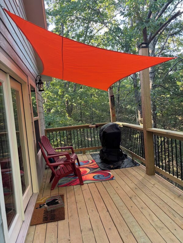 A wooden deck with a barbecue grill, two Adirondack chairs, and a colorful rug.
