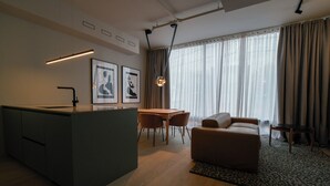 A modern living room with a sofa, a dining table with chairs, a kitchen area with a sink, and a large window with curtains.