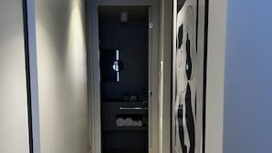 A narrow hallway with a wooden floor, a door leading to a bathroom, and a wall with a mirror and framed artwork.
