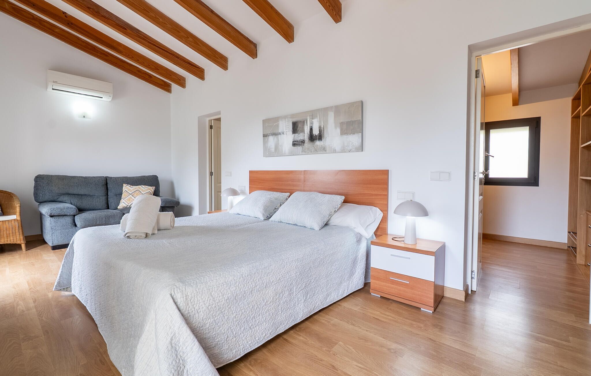 A bedroom with a large bed, wooden ceiling beams, a grey sofa, and a bedside table with a lamp.