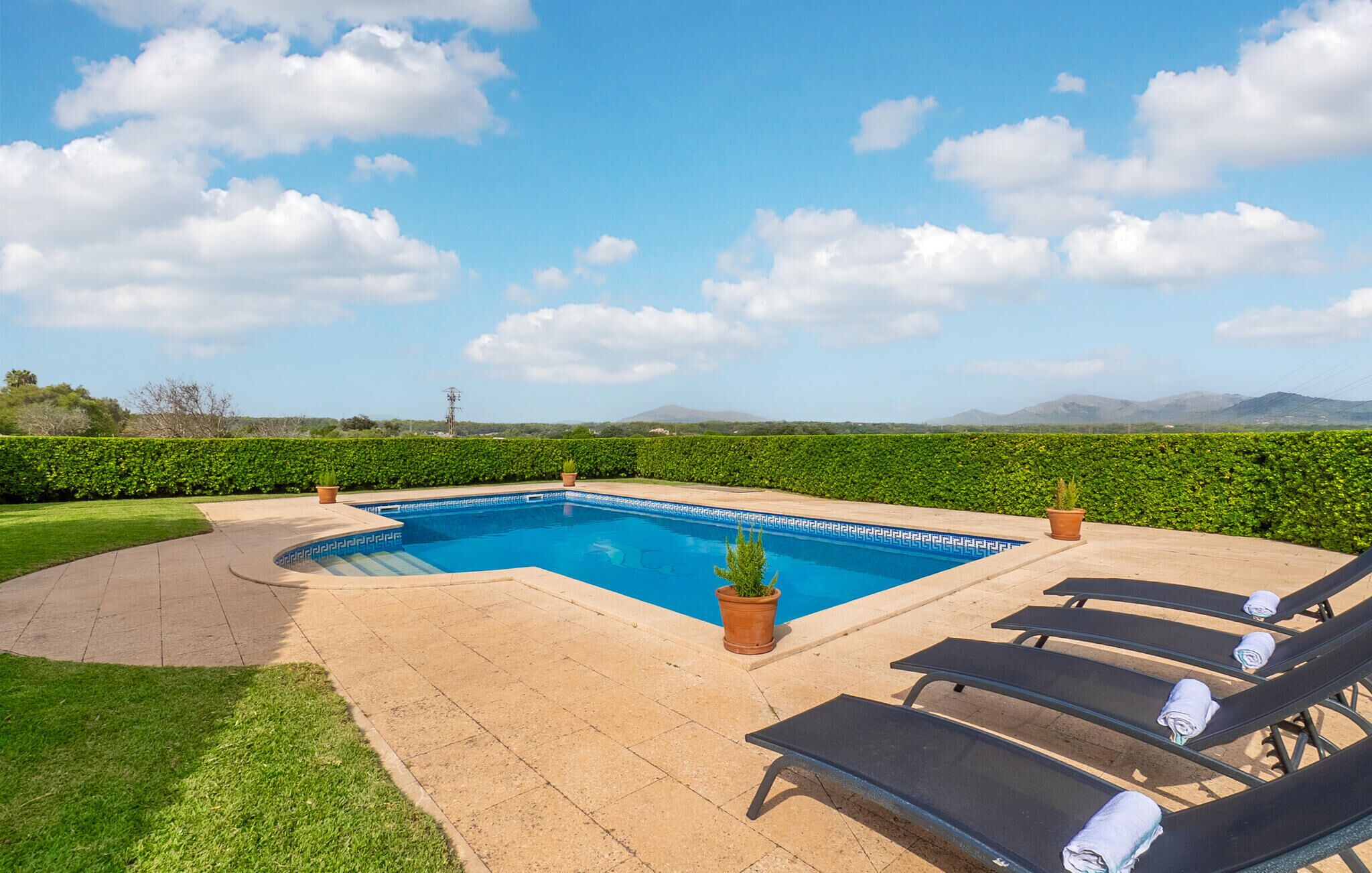 Piscina com padrão geométrico de azulejos, cercada por um deck pavimentado e área gramada. Há espreguiçadeiras e plantas em vasos.