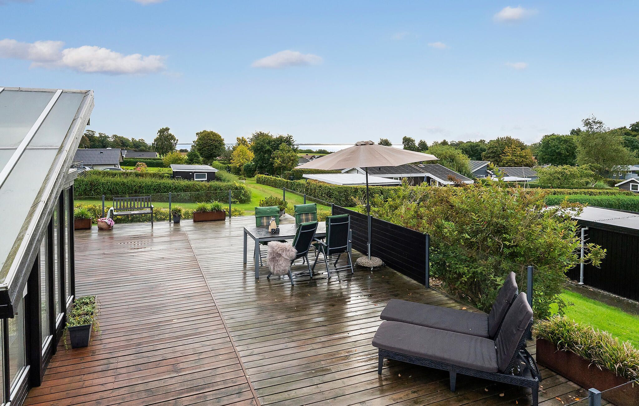 Una terraza de madera con mobiliario exterior, vista a un jardín ajardinado y una casa al fondo.