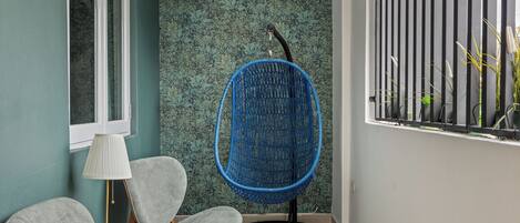 Une pièce aux murs de couleur turquoise, une fenêtre avec des stores verticaux, un fauteuil suspendu bleu, deux fauteuils gris, une petite table en bois et un lampadaire.