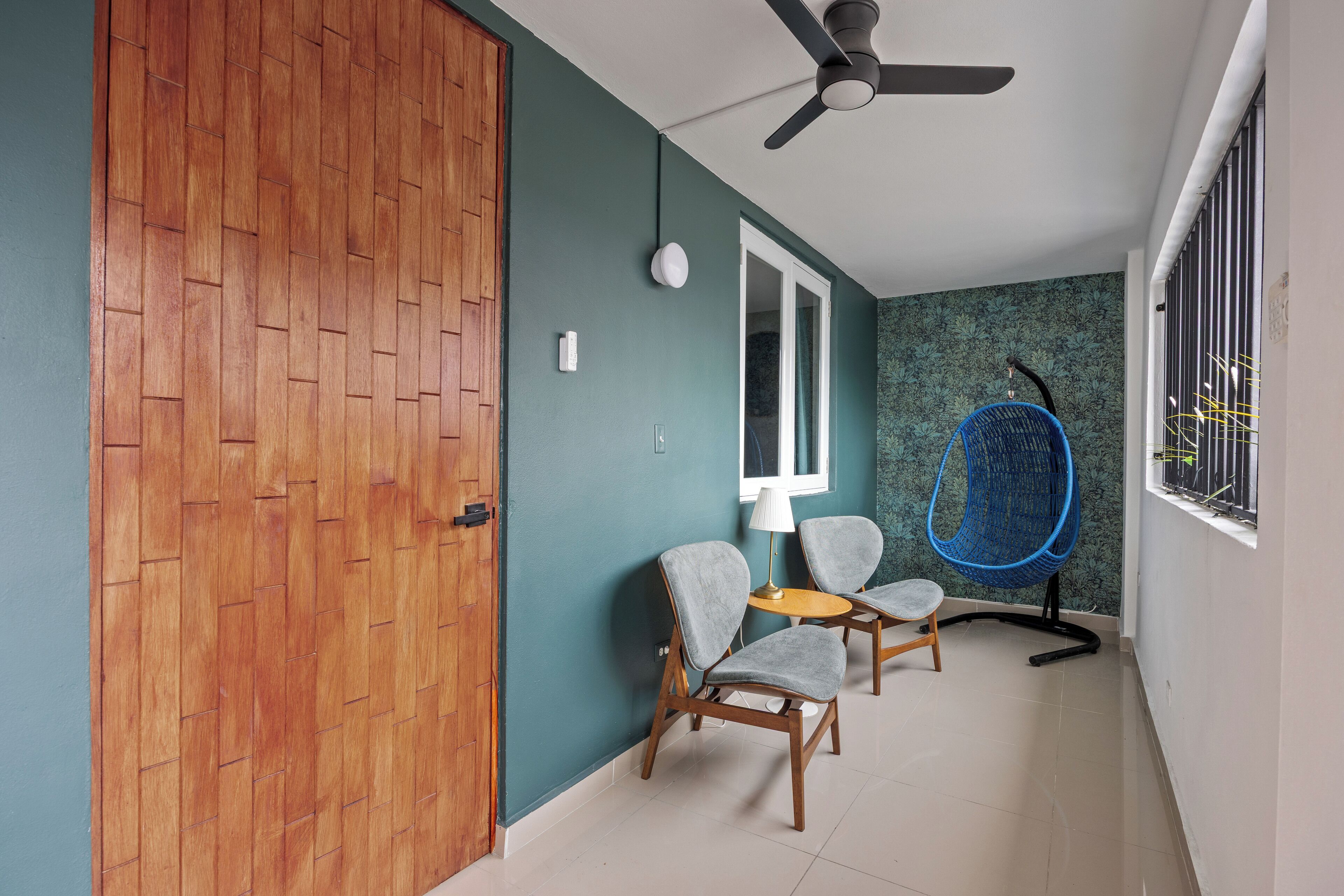 A room with a wooden door, teal walls, a ceiling fan, two grey chairs, a small round table, and a window with a metal railing.