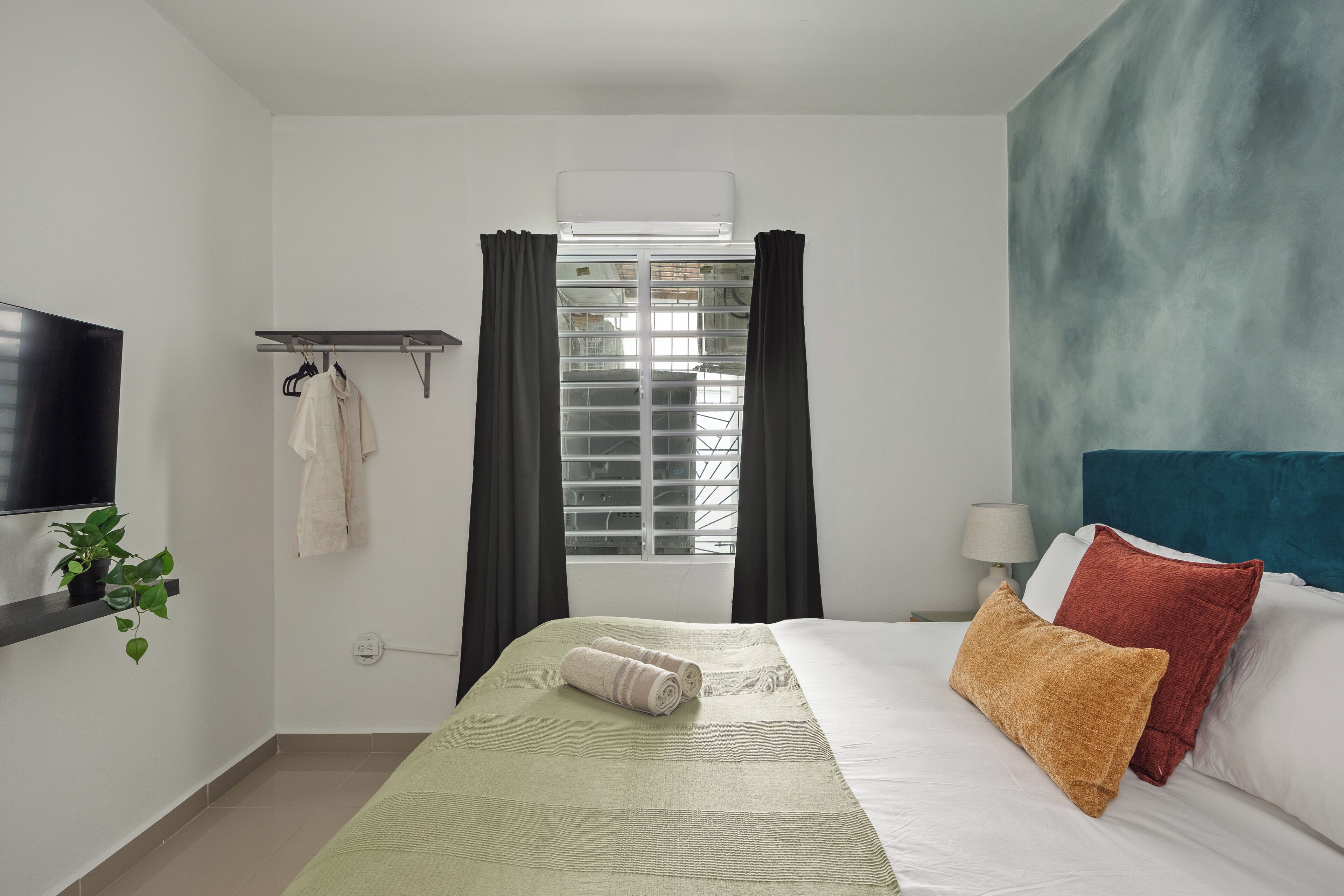 Habitación de hotel con cama, televisor, ventana con cortinas, una planta y estante en la pared.