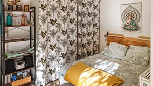Un dormitorio con una cama, una mesita de noche, un estante con libros y una lámpara, y una cortina con motivos tropicales.