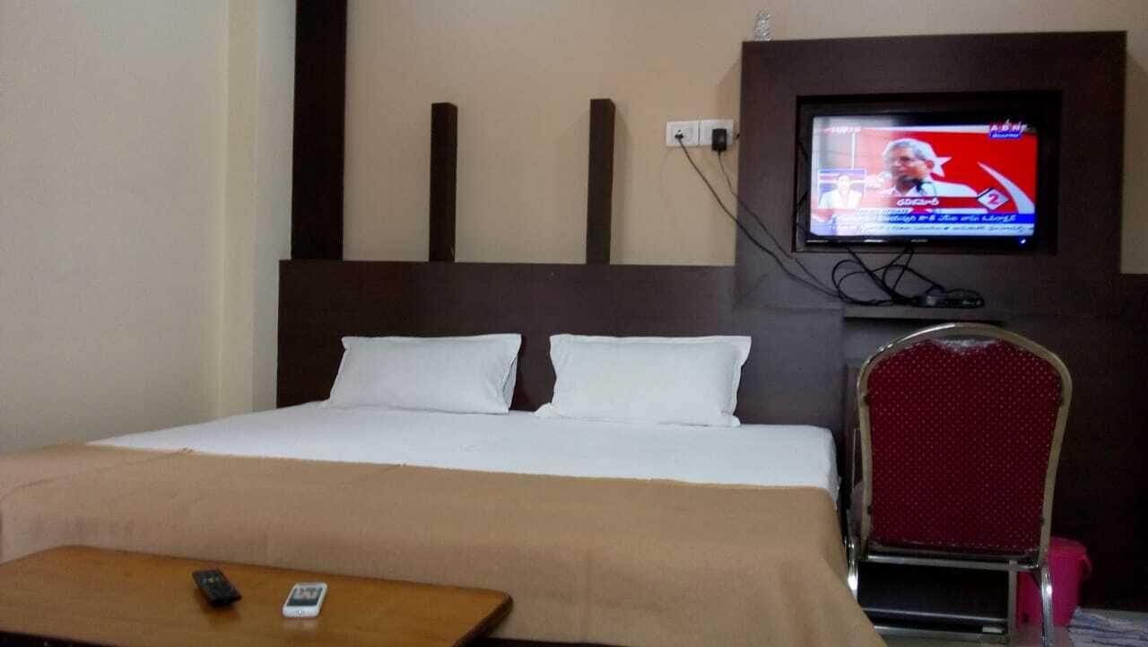 A hotel room with a bed, a television mounted on the wall, a red chair, and a wooden table with a remote control.