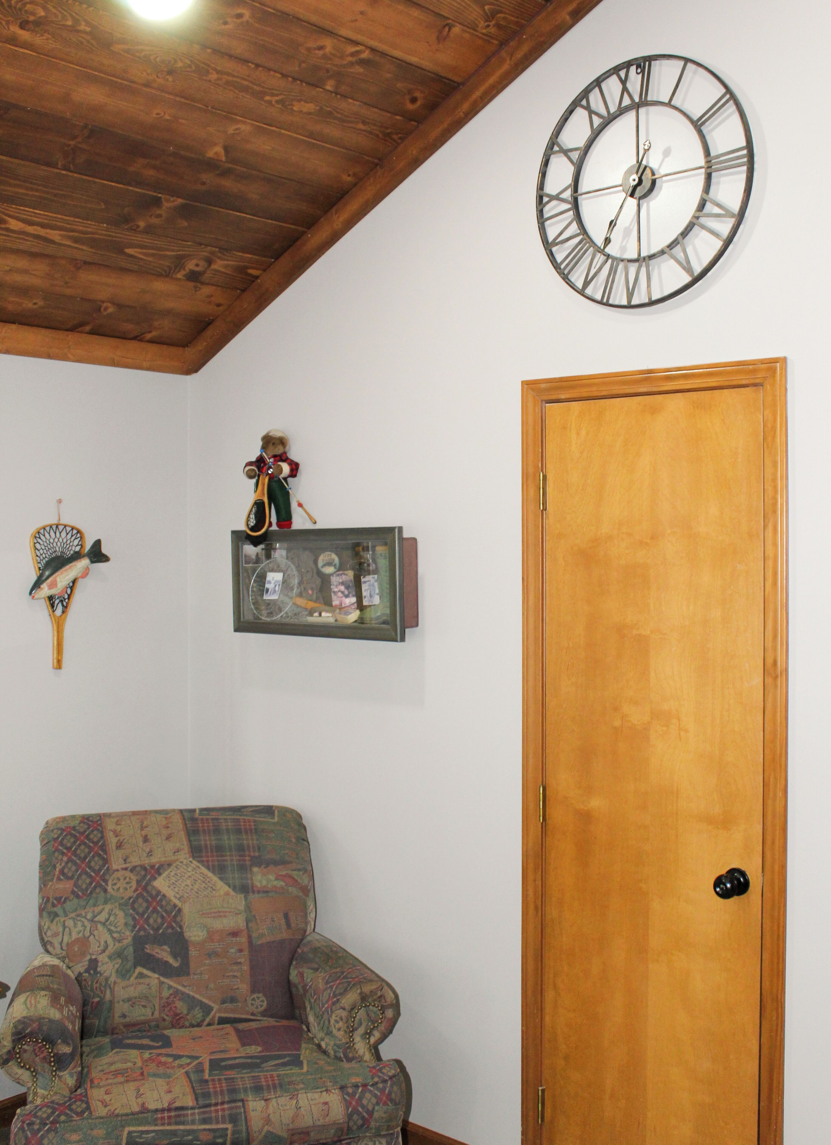 A room with a wooden door, a patterned armchair, a wall clock, and a shelf with various items.