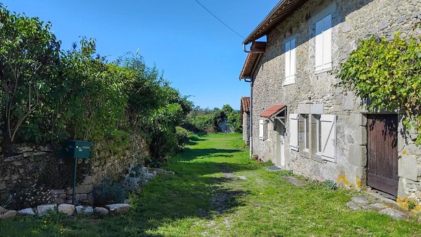 A narrow stone pathway leading to a rustic stone building with a red-tiled roof and white shutters.