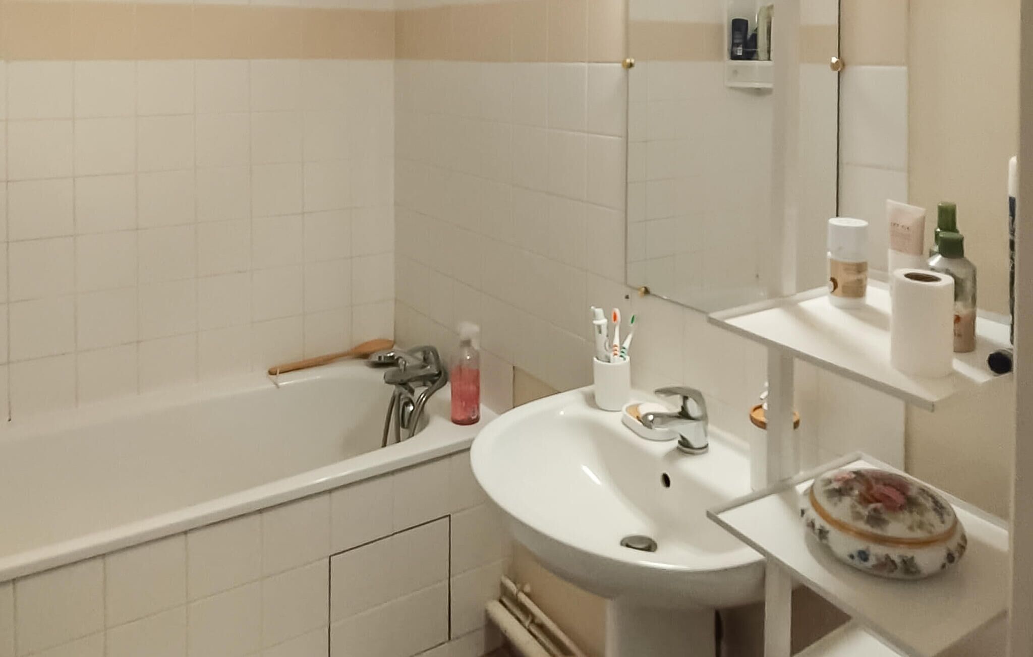 Un baño con bañera, lavamanos y espejo. Hay artículos de aseo en el lavamanos y un dispensador de jabón en la pared.