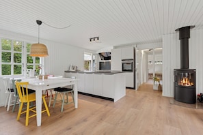 Een moderne keuken met witte kasten, een centraal eiland, een eethoek met gele stoelen en een houtgestookte kachel.