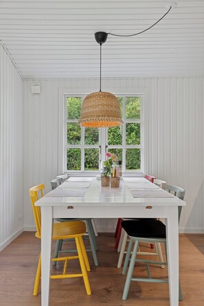 A dining area with a white table, yellow and green chairs, a hanging basket light, and a window with greenery outside.