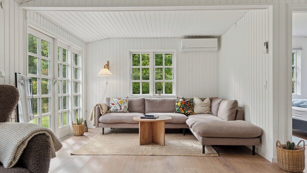 A spacious living room with a large sectional sofa, a round wooden coffee table, and a large window with a view of greenery. - Renovated classic cottage close to beach (Roslev)