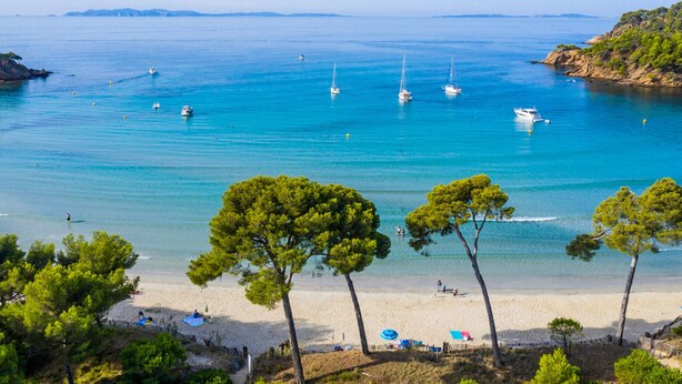 A beach with clear turquoise water, several sailboats, and a sandy shore with trees.