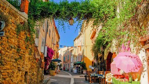 A narrow street lined with buildings, some with balconies and greenery. There are tables and chairs outside a shop, and a cart with items.