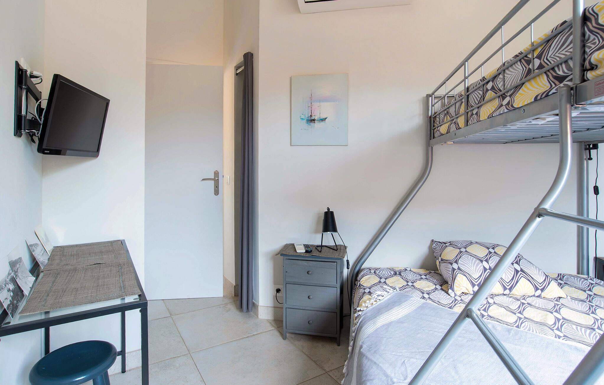 Une chambre compacte comprenant un lit superposé, un bureau avec une chaise, une petite table de chevet avec une lampe et un téléviseur fixé au mur.