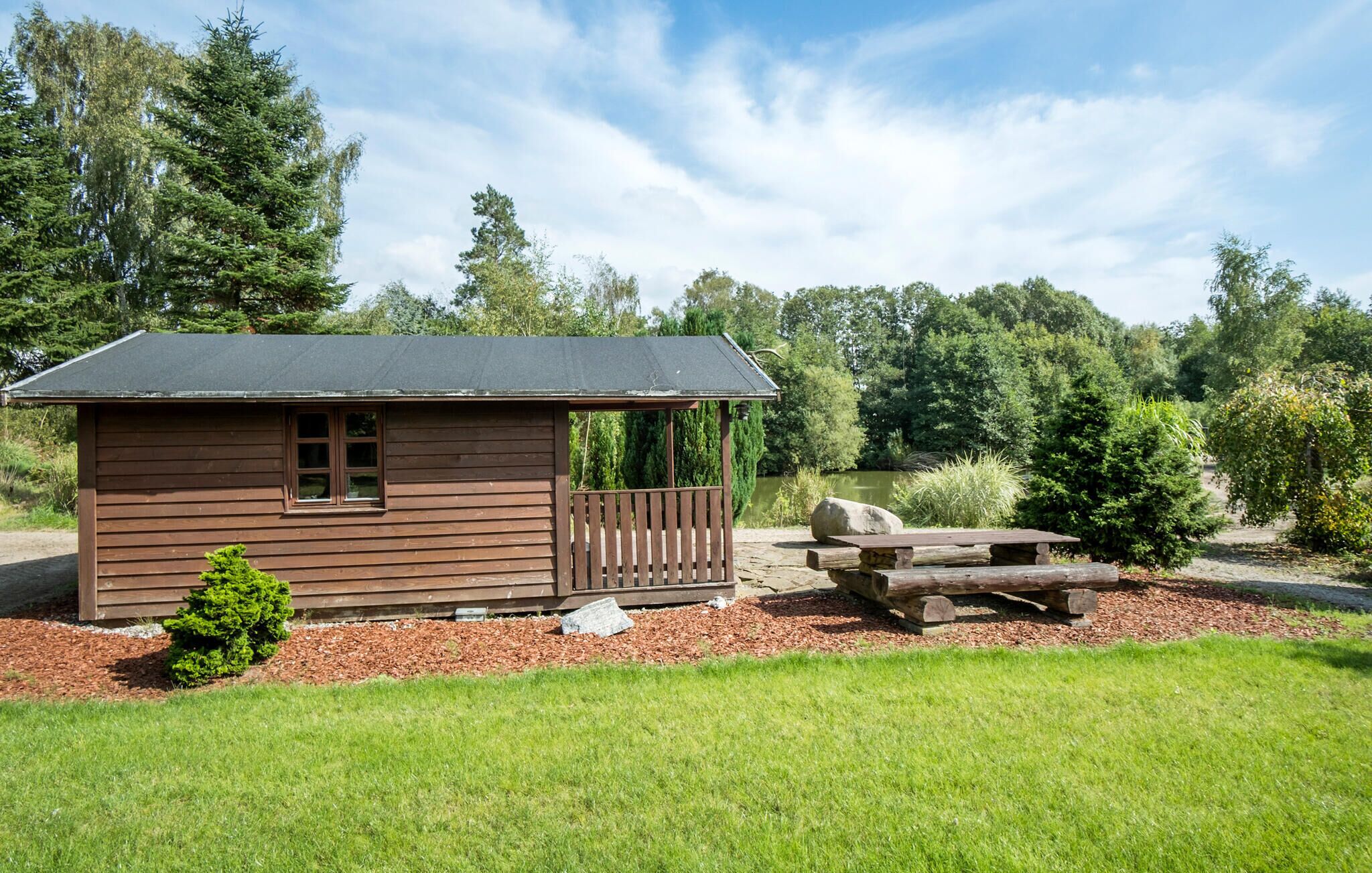 Eine kleine Holzhütte mit Veranda und Picknicktisch, umgeben von Wiese und Bäumen.