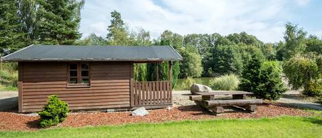 Eine kleine Holzhütte mit Veranda und Picknicktisch, umgeben von Wiese und Bäumen.