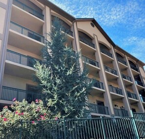 A multi-story building with balconies and a large evergreen tree in the foreground. - Smoky Bear Crossing - Downtown Corner Condo, Walk to Attractions (Gatlinburg)