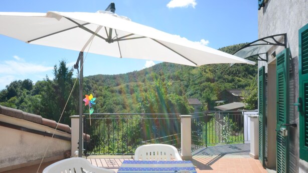 A balcony with a table and chairs, a white umbrella, and a view of a green hillside.