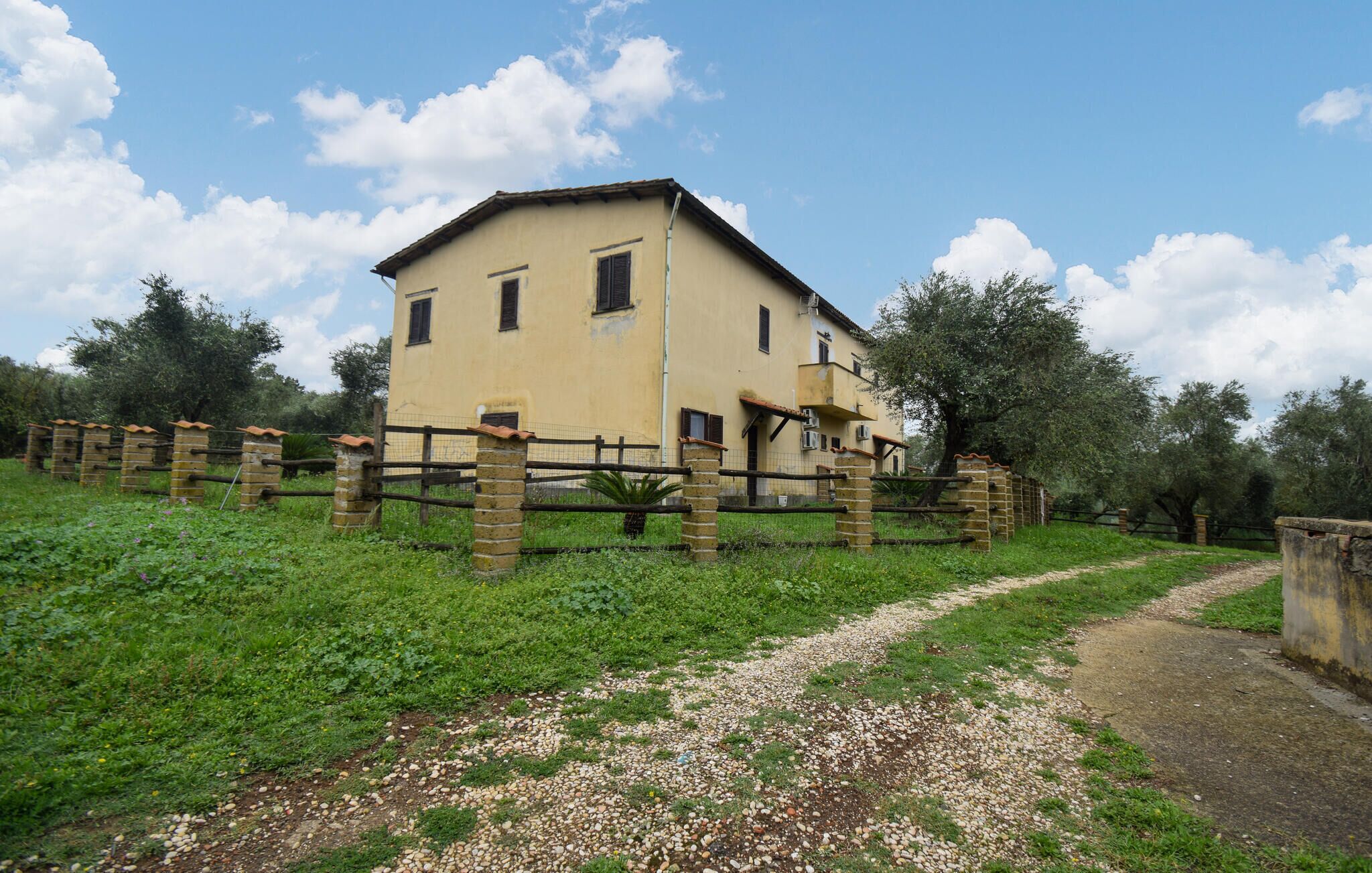 A two-story building with a yellow facade, surrounded by a stone fence and a gravel path leading to it.