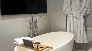 Une salle de bain moderne avec une baignoire sur pieds, un téléviseur fixé au mur et deux peignoirs blancs suspendus à des crochets.