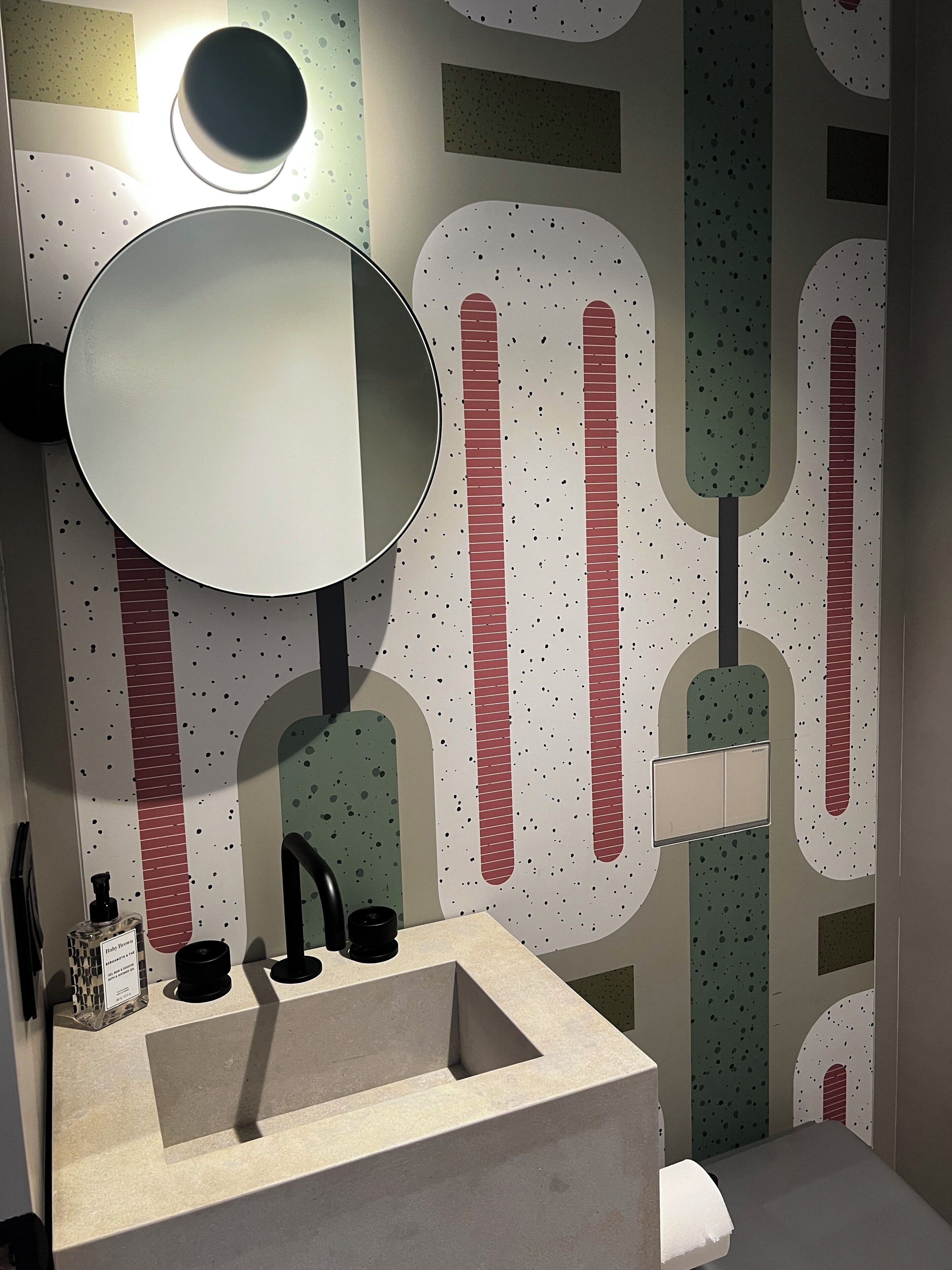 A bathroom with a round mirror, a sink, and a toilet. The walls have a speckled pattern with abstract shapes.