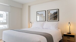 Una cama bien hecha con sábanas blancas y una manta gris, una mesita de noche con lámpara, y dos cuadros en la pared.