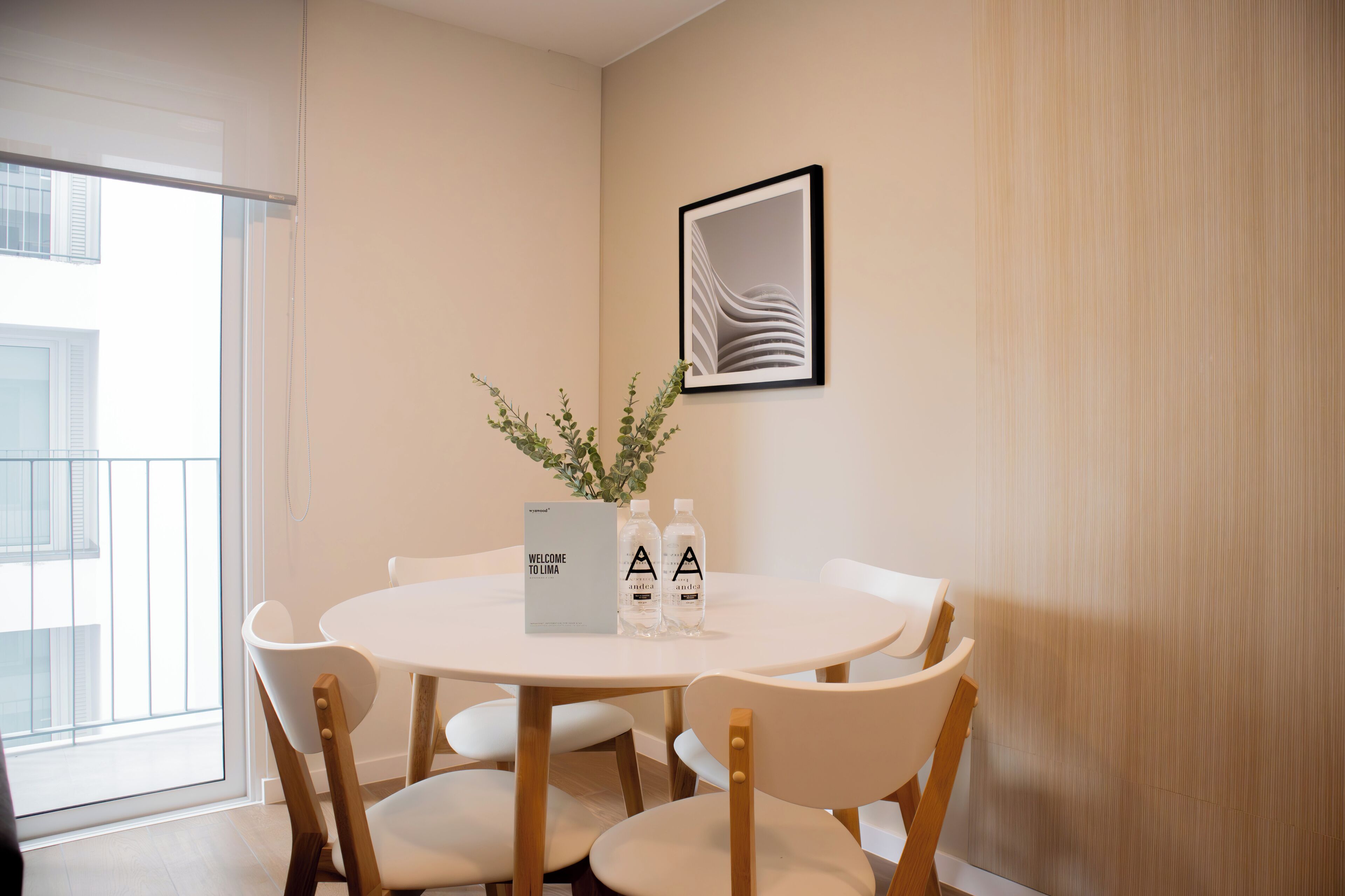 A modern dining area with a round table, white chairs, a framed picture, and a bottle of water.