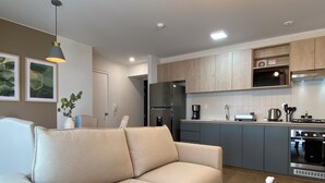 Una sala moderna con un sofá color beige, una mesa de centro redonda de madera y una zona de cocina con armarios grises y electrodomésticos de acero inoxidable.