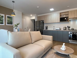 Un salon moderne avec un canapé beige, une table basse ronde en bois et un coin cuisine doté de meubles gris et d’appareils électroménagers en acier inoxydable.